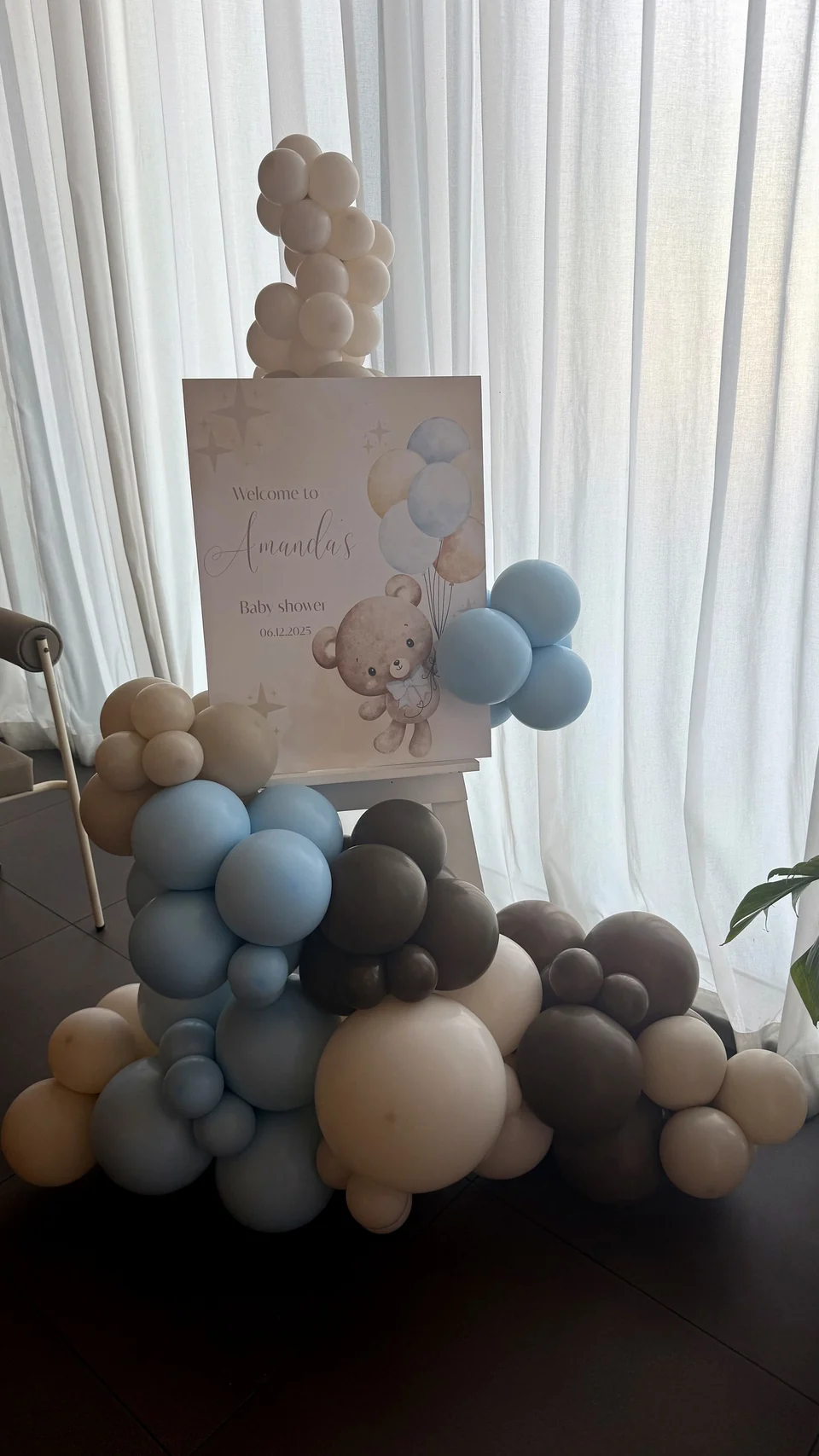 Baby shower welcome sign with a teddy bear, framed by a blue, taupe and cream balloon garland