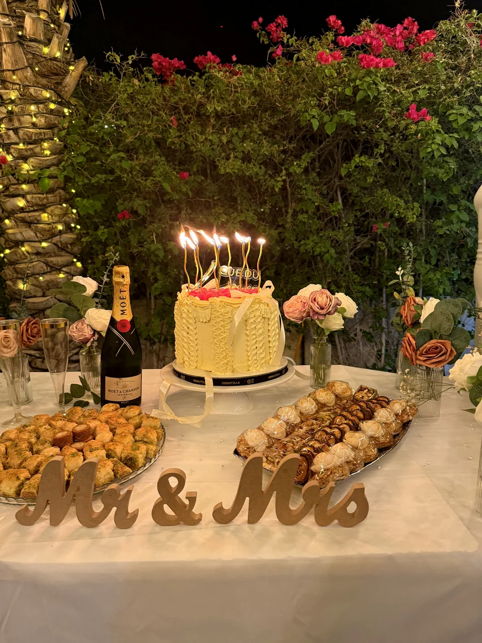 Celebration cake with lit candles beside a gold Mr and Mrs sign, champagne and pastries
